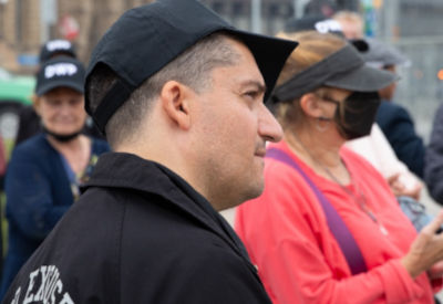 A side profile of a man wearing a black cap with "DWP" written on it, standing in a crowd. People in the background are casually dressed, some wearing face masks.