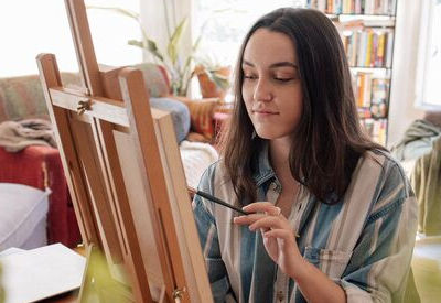 A young woman seated indoors painting on an easel. She is focused, wearing a striped shirt, with bookshelves and cozy furnishings in the background.