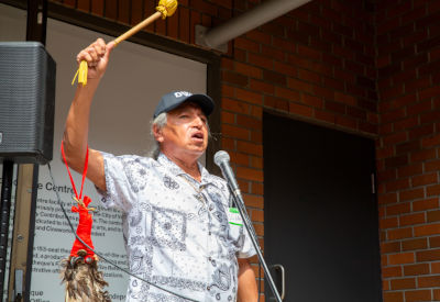 An older man speaking passionately into a microphone while raising a drum stick.