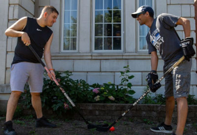 Two individuals are playing street hockey outside a building with large windows. Both hold hockey sticks and smile as they engage in the game, surrounded by greenery.