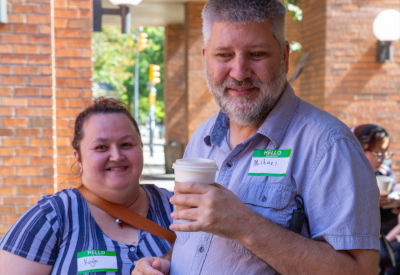 A man and woman smile at the camera while holding coffee cups at an outdoor gathering. Both wear name tags that read "Kait" and "Michael."