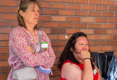 Two women stand against a brick wall, attentively listening during an event. One woman wears a floral blouse and crosses her arms, while the other rests her hand on her chin. Both wear name tags.