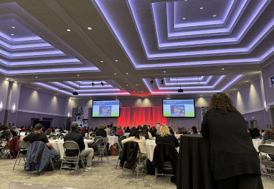 A large conference room filled with people sitting at round tables while a presentation is displayed on two large screens at the front; the room is illuminated with modern, recessed lighting that creates a professional and vibrant atmosphere.