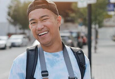 A cheerful young man wearing a backward baseball cap and a tie-dye shirt, carrying a camera around his neck, smiling brightly on a busy street; the background includes blurred vehicles and urban surroundings, giving a lively and dynamic feel to the image.