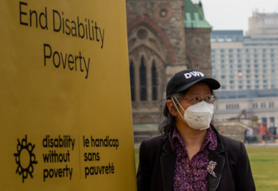 A person wearing a black "DWP" hat and a face mask stands beside a tall, bright yellow banner that reads "End Disability Poverty" alongside the logos for Disability Without Poverty and its French translation; in the background, the historic architecture of Parliament Hill in Ottawa can be seen, lending a sense of purpose and activism to the scene.