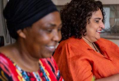 Two women sitting at a kitchen table, smiling and enjoying a conversation. The woman in the foreground wears a colorful patterned dress and a black headwrap, while the woman in the background wears an orange top and curly hair.