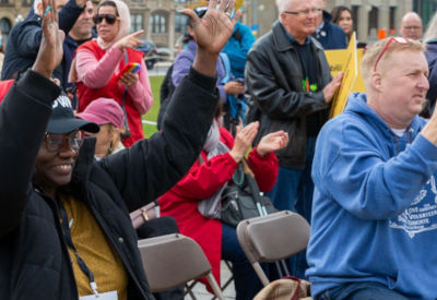 A group of people seated outdoors at an event, applauding and raising their hands. A woman in a black jacket and cap with 'DWP' written on it is smiling and engaging with the activity. The atmosphere is lively and supportive.