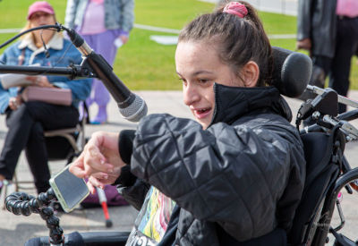 A woman seated in a wheelchair speaks into a microphone, smiling and gesturing expressively. She wears a black quilted jacket and has her hair tied back with a pink scrunchie. The background shows an outdoor event setting with other participants seated.