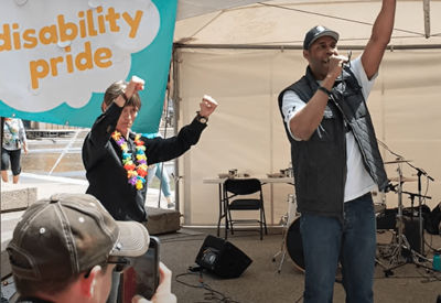 Two individuals on stage at a Disability Pride event, both raising their arms enthusiastically. A banner reading 'Disability Pride' is visible in the background, along with a small musical setup.