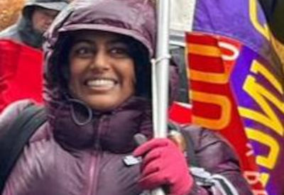 A woman in a purple jacket and hood, smiling and holding a brightly colored union flag during a rally on a rainy day.