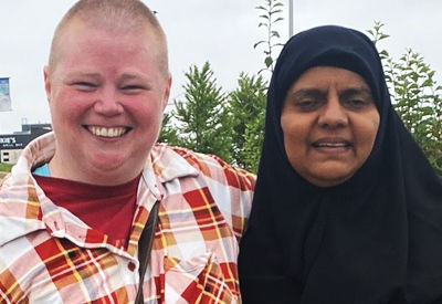 Two women sitting side-by-side in a kitchen. One is wearing a black headscarf and a vibrant patterned top, smiling slightly. The other woman, with curly hair and wearing an orange blouse, is laughing. Both appear to be enjoying a conversation or activity together.