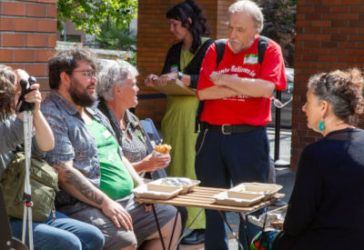 A small group of people gathered around a table in an outdoor setting. They engage in conversation while eating from compostable trays. One man in a red T-shirt leans casually against the table.