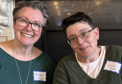 Two smiling women sitting side by side indoors. They wear name tags reading "Meaghen" and "Julia." Both are dressed casually in green tops and glasses, exuding a warm, friendly vibe.