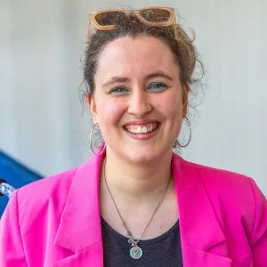 Elizabeth Lowe, wearing a bright pink blazer, a gray top, and a silver pendant necklace, smiles warmly at the camera. Her hair is tied back, and she wears sunglasses on her head, with hoop earrings and teal eyeshadow in a well-lit indoor setting.