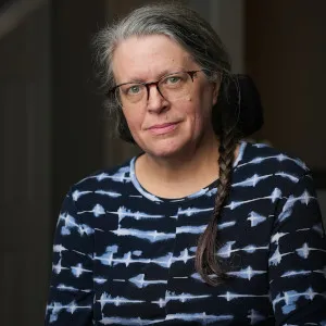 Michelle Hewitt, wearing glasses and a dark patterned top with her hair in a braid, looks calmly at the camera in a softly lit indoor setting.