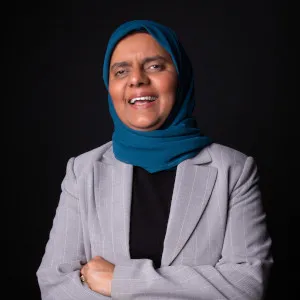 Rabia Khedr, wearing a teal hijab and a light gray blazer over a black top, smiles warmly with her arms crossed, set against a black background.