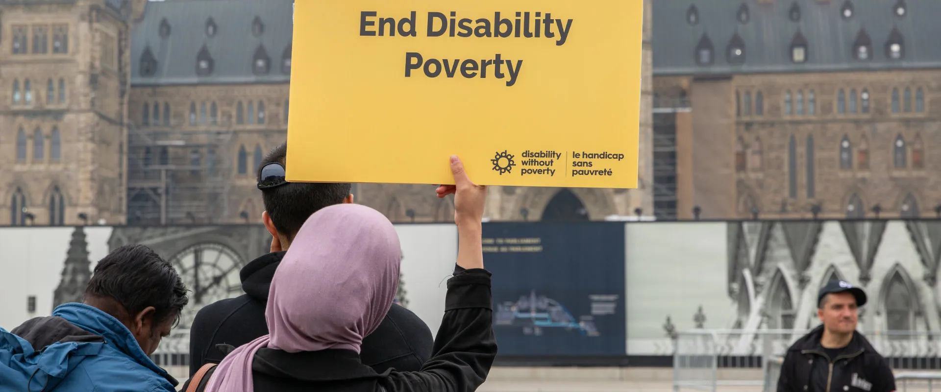 A group of people participating in a demonstration, with one individual prominently holding up a large yellow sign that reads 'End Disability Poverty.' The sign also features the logo and name of 'Disability Without Poverty' in both English and French. The background shows a historic building with Gothic architecture, suggesting the event is taking place in a civic or governmental area. The scene captures a moment of activism and advocacy.