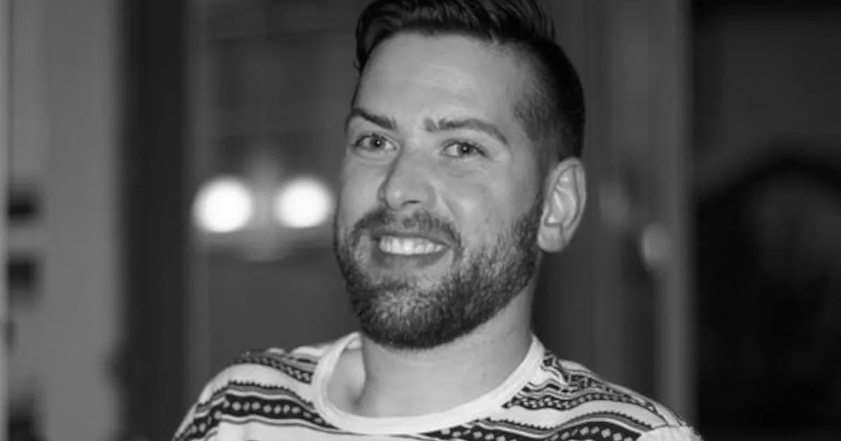 Black and white photo of a smiling man with a short beard and styled hair, wearing a patterned T-shirt. The background is softly blurred, with hints of lights and furniture, suggesting an indoor setting.