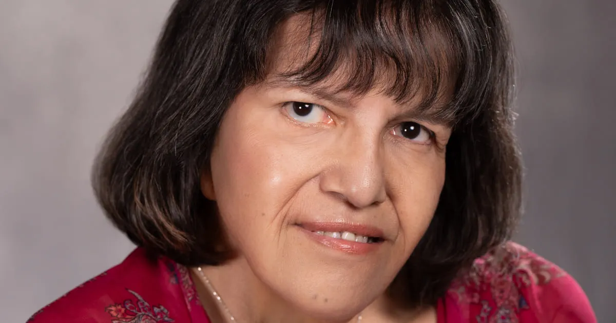 A close-up portrait of Sandra Pronteau with short, dark hair and bangs, wearing a red top with floral patterns, looking directly at the camera with a slight smile. The background is softly blurred in shades of gray.