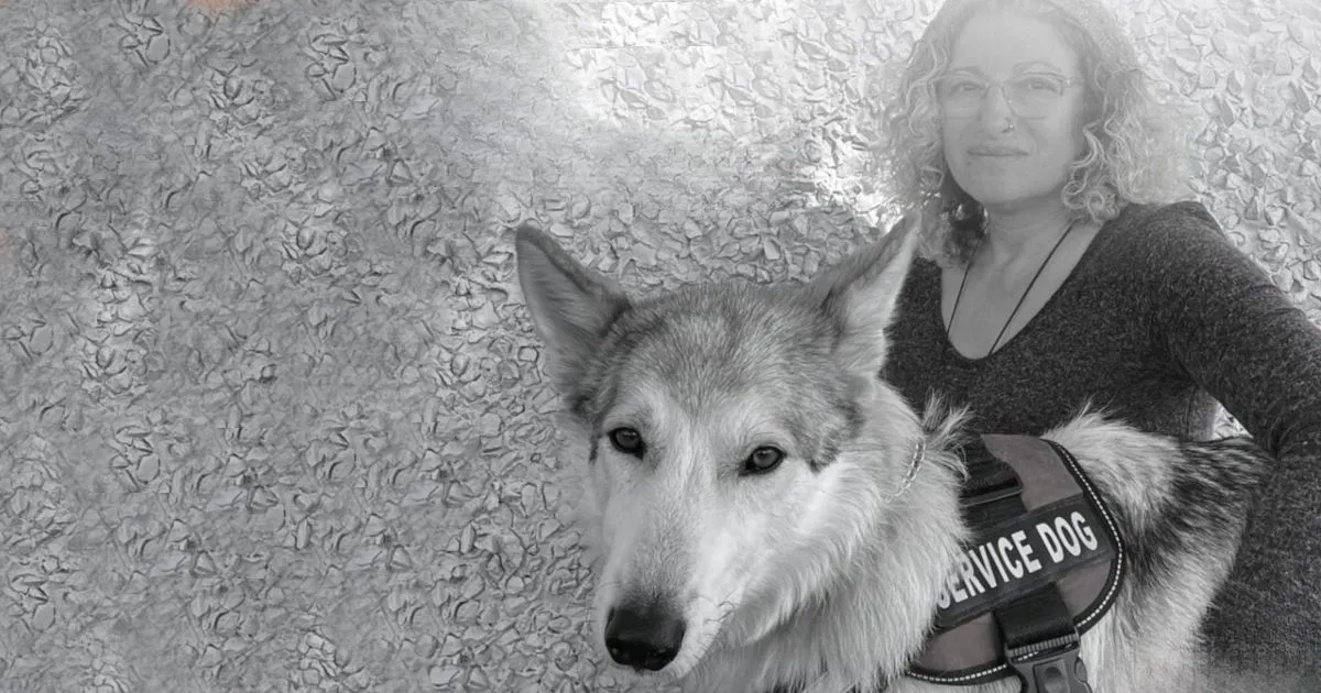 Black and white image of a person with curly hair, glasses, and a calm expression, standing beside a large service dog wearing a vest labeled 'Service Dog.' The background has a textured, floral pattern, giving the photo an artistic feel.